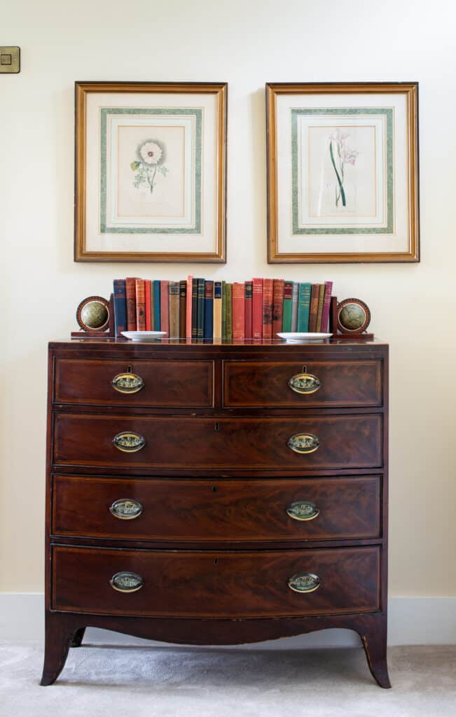 A vintage wooden dresser with ornate handles, topped with two rows of colorful books, evokes the charm of luxury travel. Two decorative globes sit at each end like worldly guides. Above, two framed botanical prints hang on a cream wall. The floor is carpeted in light gray. David Pearson Travel