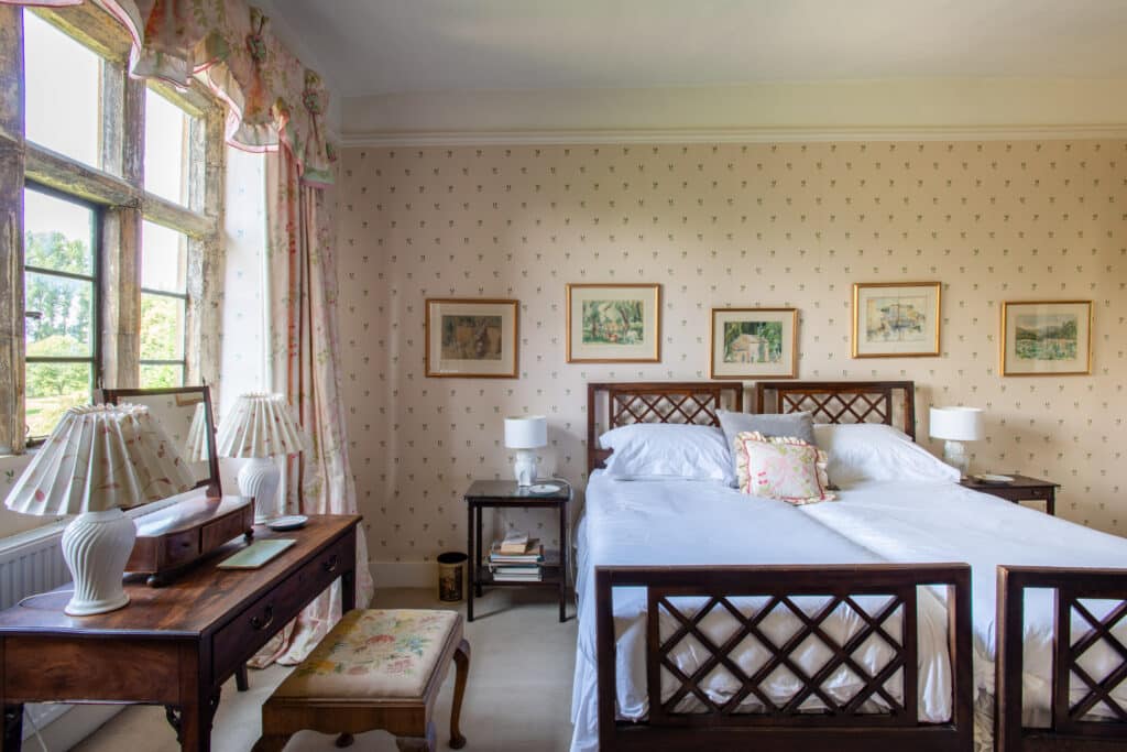Experience luxury travel in this traditional bedroom, where a large bed with white linens and wooden lattice headboards promises restful nights. Three framed pictures adorn the wall above, while a wooden dressing table with lamps sits near a window draped in floral curtains and subtle patterned wallpaper. David Pearson Travel
