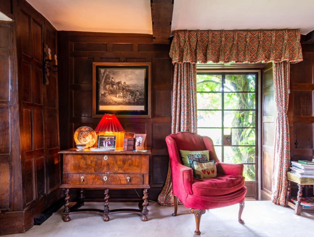 A cozy room with dark wood paneling features a red wingback chair beside a side table adorned with a lamp and framed photos, capturing memories of past luxury travels. A large window with patterned curtains overlooks lush greenery, while books and pictures are elegantly arranged throughout. David Pearson Travel
