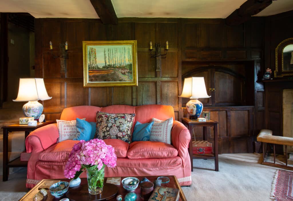 A cozy living room exuding the elegance of luxury travel, featuring a pink sofa, decorative cushions, and wooden paneling. Two lit lamps flank the sofa on side tables, while pink flowers in a vase grace the coffee table. A framed landscape painting elegantly hangs above the sofa. David Pearson Travel