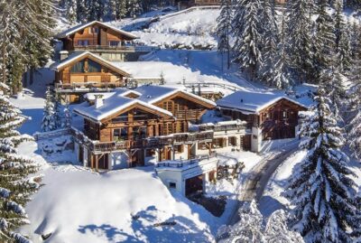 A luxury travel experience awaits at this snow-covered chalet nestled among pine trees on a hillside. The wooden building features multiple sloped roofs and balconies. A narrow path winds its way to the chalet, while the surrounding landscape is blanketed in snow under a clear blue sky. David Pearson Travel