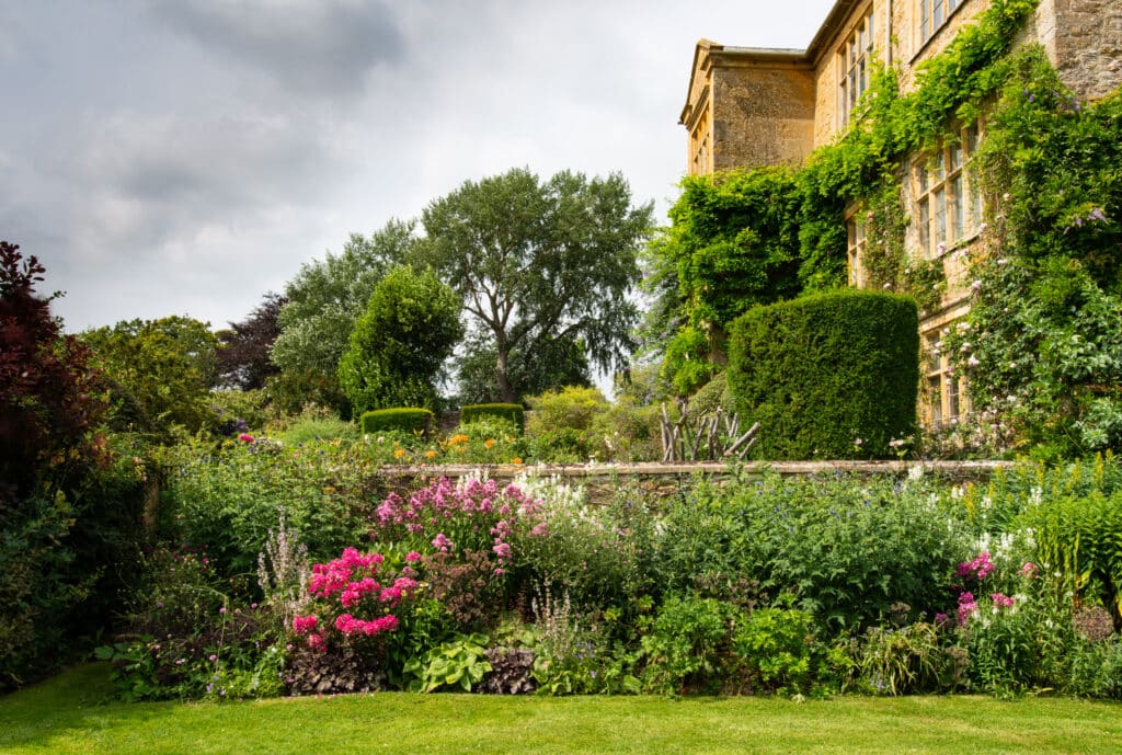 A lush garden with vibrant flowers and greenery graces the frontage of a large, ivy-covered stone house, reminiscent of a luxury travel escape. The overcast sky casts soft light over the scene, while tall trees loom majestically in the background. David Pearson Travel