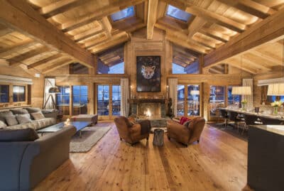 A spacious wooden chalet interior with high ceilings and large windows defines luxury travel. The room features a cozy seating area with two chairs facing a fireplace, a plush sofa, a dining table set for dinner, and a picturesque outdoor view. David Pearson Travel