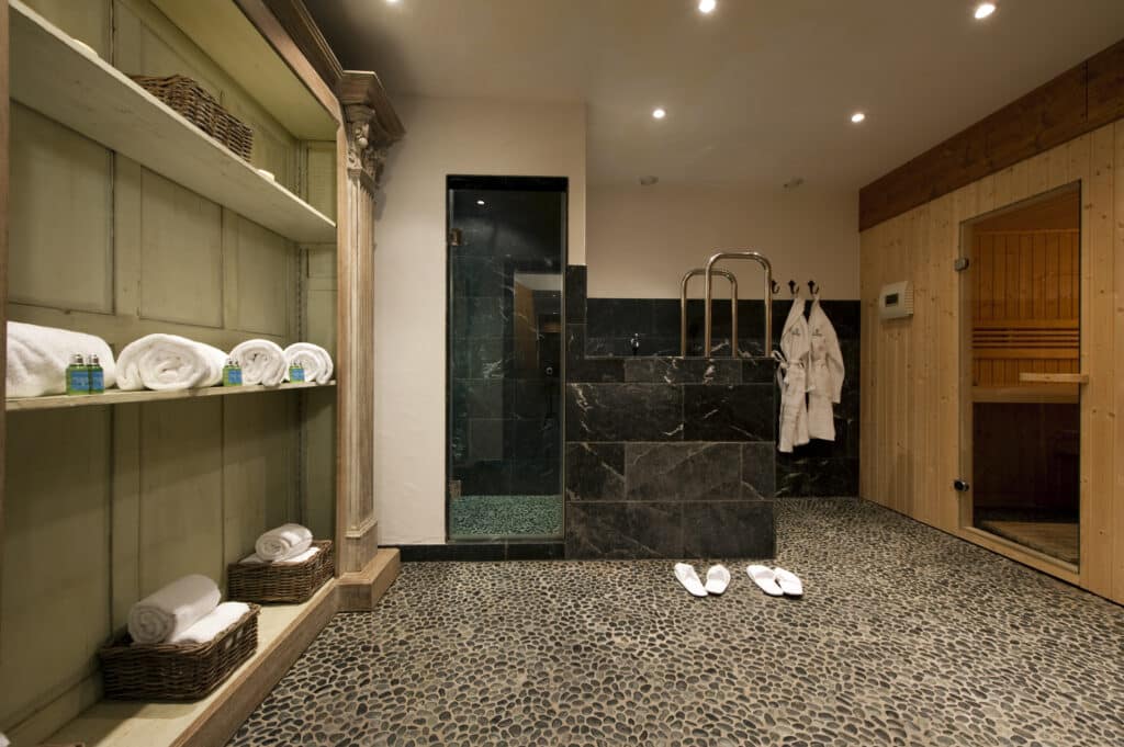 The spa room exudes luxury travel vibes with its stone-tiled floor, a shelf of rolled towels, and toiletries on the left. A glass-enclosed shower takes center stage, flanked by plush bathrobes. Slippers rest on the floor beside a wooden sauna for ultimate relaxation. David Pearson Travel