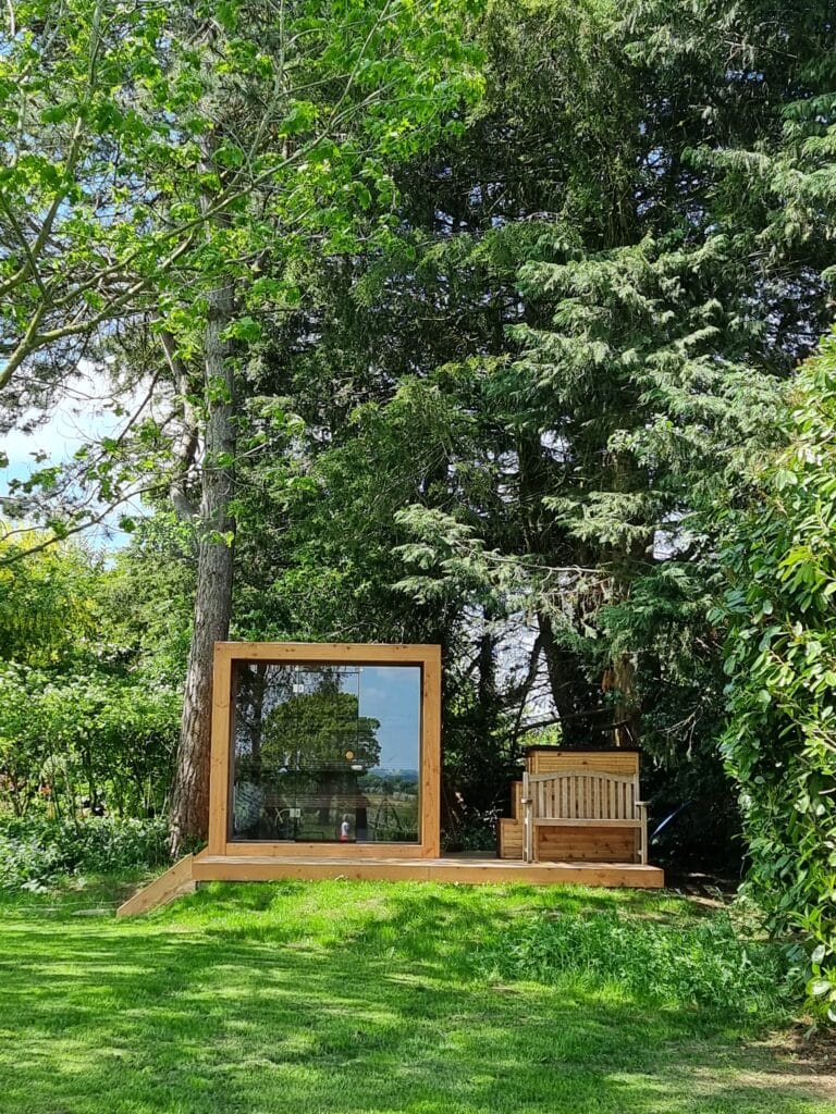 A modern wooden cabin with large glass windows sits amidst lush greenery, offering a luxury travel experience. Next to it is a wooden bench. The scene is surrounded by tall trees, creating a peaceful, natural setting as sunlight filters through the leaves. David Pearson Travel