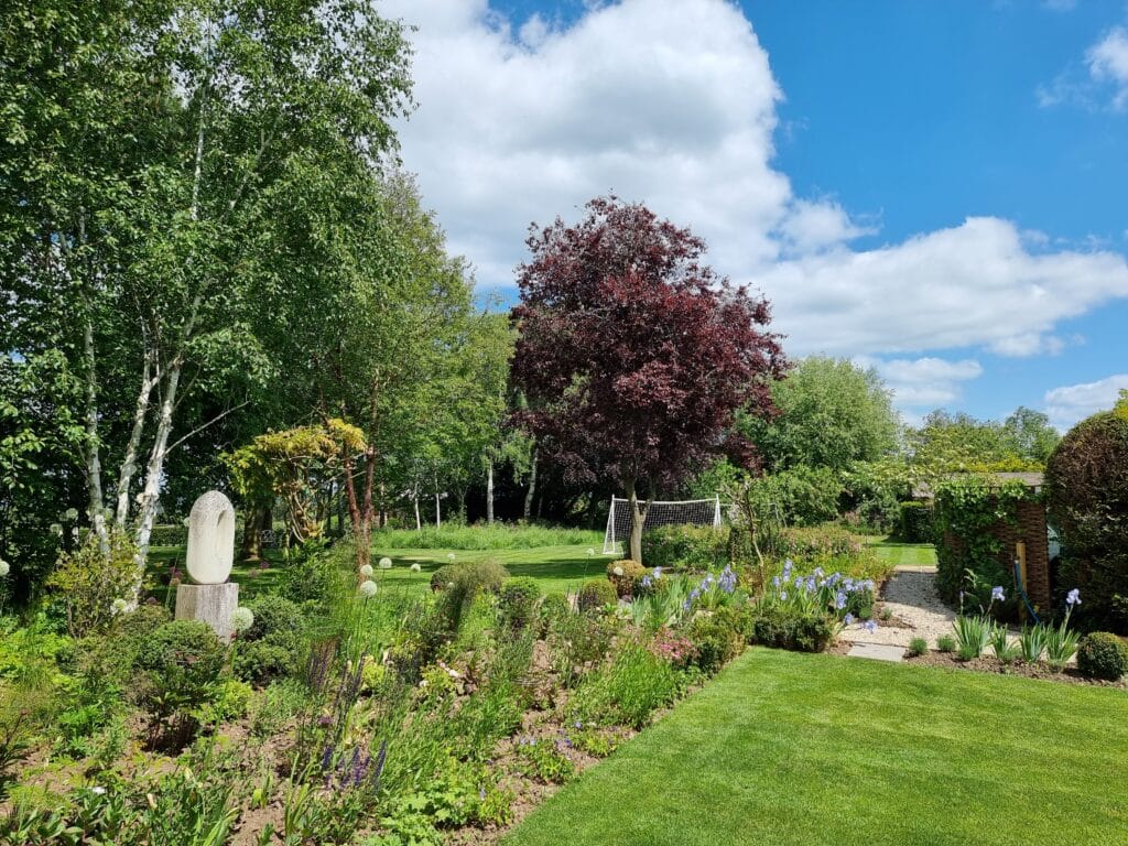 A lush garden scene reminiscent of luxury travel, with various green plants and blooming flowers. A neatly trimmed lawn is bordered by colorful flowerbeds. Tall trees with green and burgundy leaves stand against a bright blue sky dotted with fluffy white clouds. David Pearson Travel
