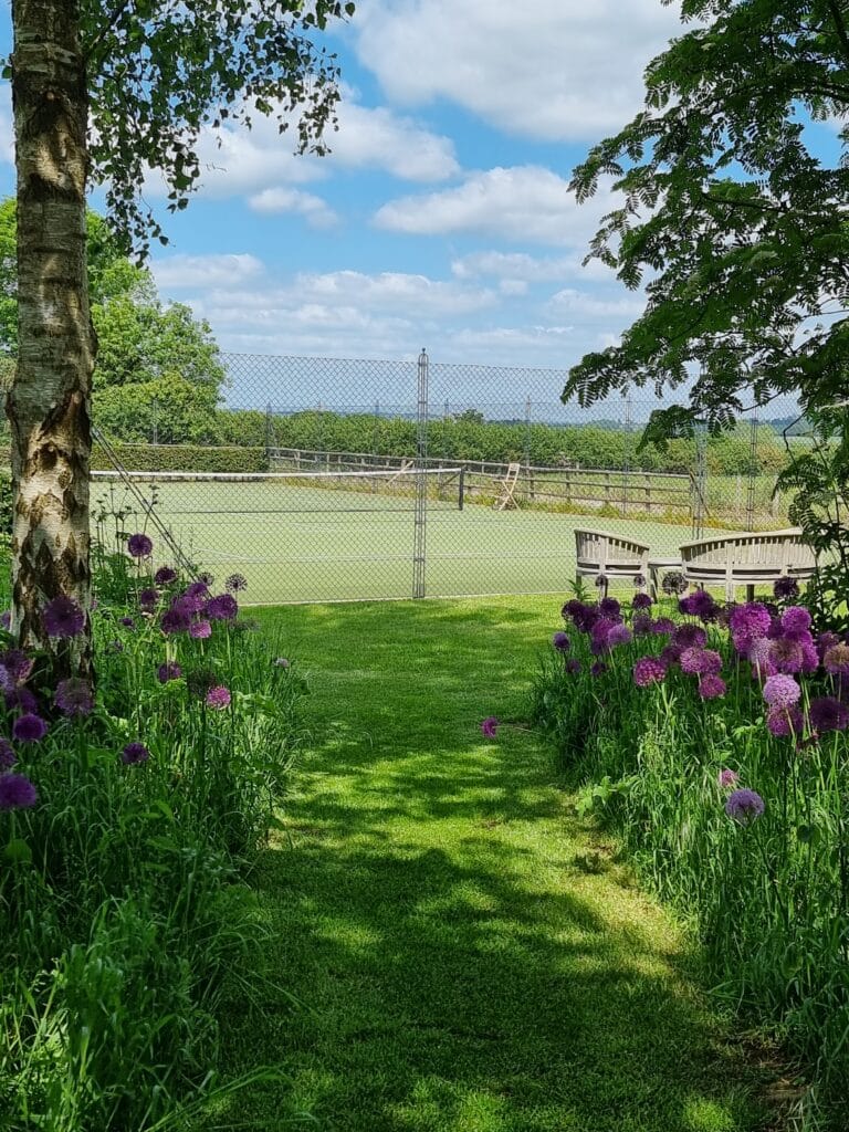 A garden path lined with tall purple allium flowers leads to a fenced pond under a partly cloudy sky, offering a touch of luxury travel right at home. Wooden benches on the right, surrounded by lush greenery and trees, create an enchanting and serene outdoor setting. David Pearson Travel