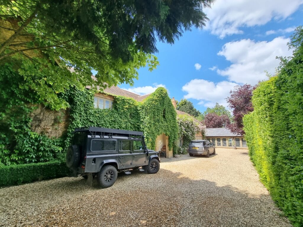 A rustic house draped in green ivy nestles amidst the trees, exuding a sense of luxury travel. Two SUVs are parked on the gravel driveway, ready for adventure. Above, the sky is bright blue with scattered clouds. David Pearson Travel