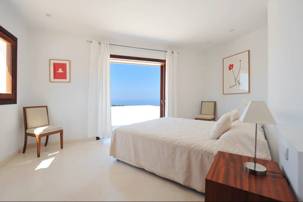 A bright bedroom with white walls offers a sense of luxury travel, featuring a double bed, wooden nightstand with a lamp, and two chairs. Large windows and an open door reveal views of the blue sky and sea. Two framed artworks grace the walls, as parted curtains allow natural light in. David Pearson Travel