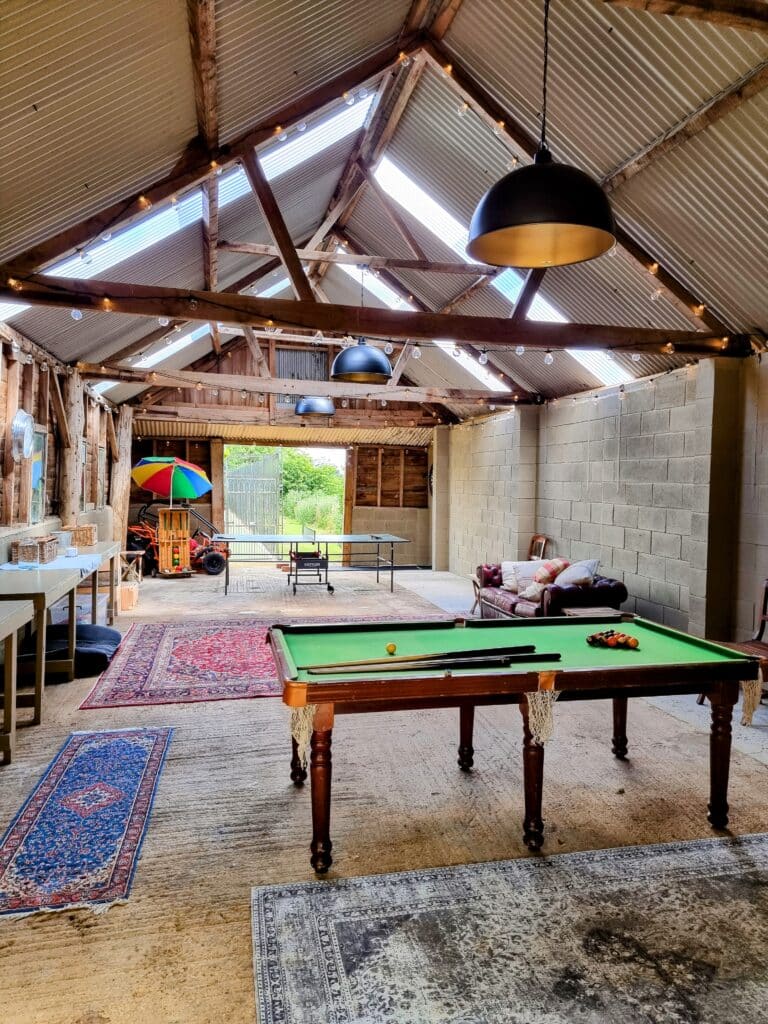 Spacious barn interior with exposed beams, embodying a touch of luxury travel. A pool table sits at the center, while a ping pong table and sofa rest in the background. Colorful umbrellas and rugs enhance the decor, as large pendant lights elegantly illuminate the space. David Pearson Travel