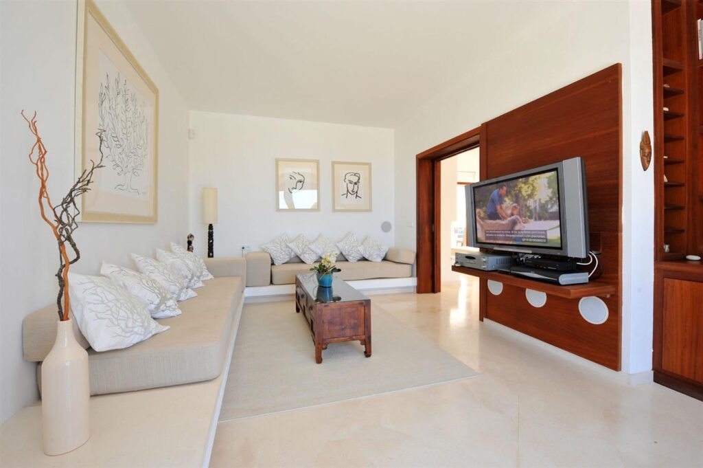 A modern living room exudes a sense of luxury travel, with beige couches adorned by white pillows reminiscent of first-class comfort. A wooden coffee table rests on a light rug, while abstract artwork graces the walls. The flat-screen TV mounted on a wooden panel completes this minimalist oasis. David Pearson Travel