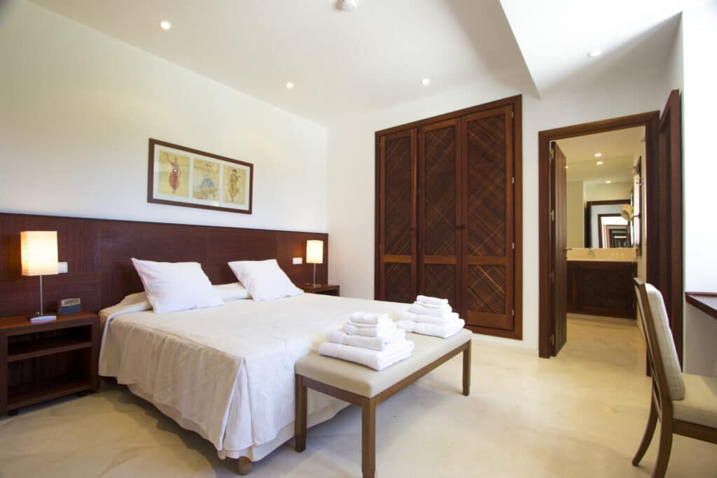 A modern bedroom reminiscent of luxury travel features a large bed with white bedding and a bench at the foot. Twin bedside tables with lamps flank the bed, while a wooden wardrobe stands nearby. An open door leads to the bathroom, and framed artwork adds sophistication above the bed. David Pearson Travel