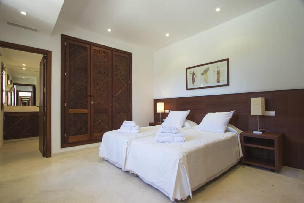 A hotel room with two single beds pushed together, each draped in white linens and towels. The wooden furniture complements a large closed wardrobe, while a bathroom is visible through an open door. Two framed artworks above the beds add a touch of luxury travel charm. David Pearson Travel