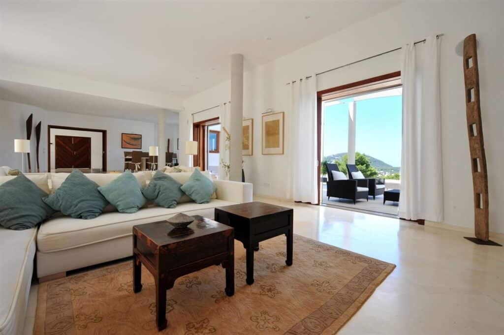 The spacious living room exudes luxury travel vibes, featuring white sofas adorned with teal pillows. A wooden coffee table rests on a beige patterned rug. Glass doors invite you to a balcony with outdoor seating, offering views of lush greenery and hills, complemented by modern art and a large sculpture. David Pearson Travel