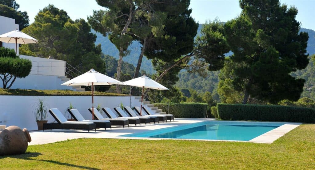 A serene outdoor setting with a rectangular swimming pool surrounded by sun loungers and umbrellas. This luxury travel destination boasts lush greenery and tall trees, with mountains visible in the background under a clear sky, offering a perfect escape into natures elegance. David Pearson Travel