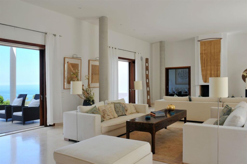 Modern living room with white sofas, a wooden coffee table, and floor-to-ceiling windows. Beige curtains and decor enhance the neutral tones. The space reflects luxury travel vibes with an outdoor seating area and ocean view visible through the open sliding glass doors. David Pearson Travel