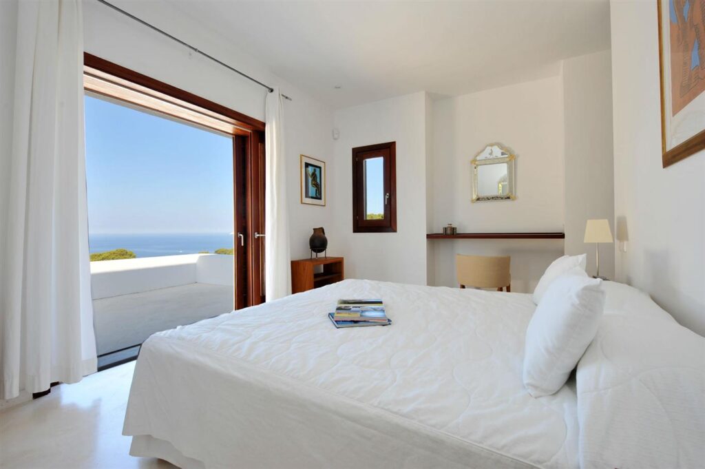 The bright bedroom, ideal for luxury travel, boasts a large open window with a stunning sea view. Featuring a white bed, light-colored walls, a small desk with a chair, and a wall mirror. A stack of books lies on the bed, while light curtains elegantly frame the window. David Pearson Travel