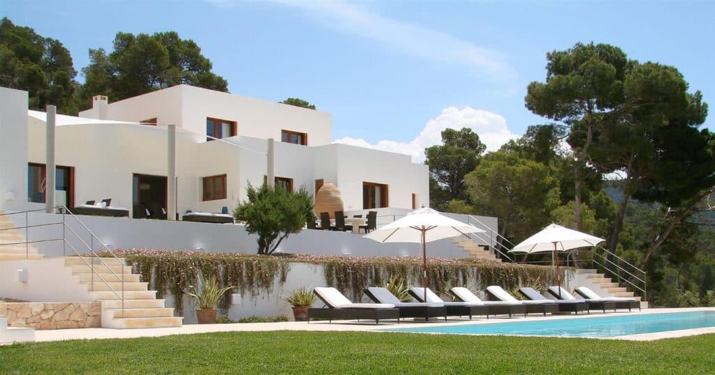 The image showcases a modern white villa, ideal for luxury travel, with multiple levels nestled among trees. It features a swimming pool and a row of sun loungers with umbrellas on a grassy area in the foreground. Steps lead up to the house, epitomizing elegance and relaxation. David Pearson Travel