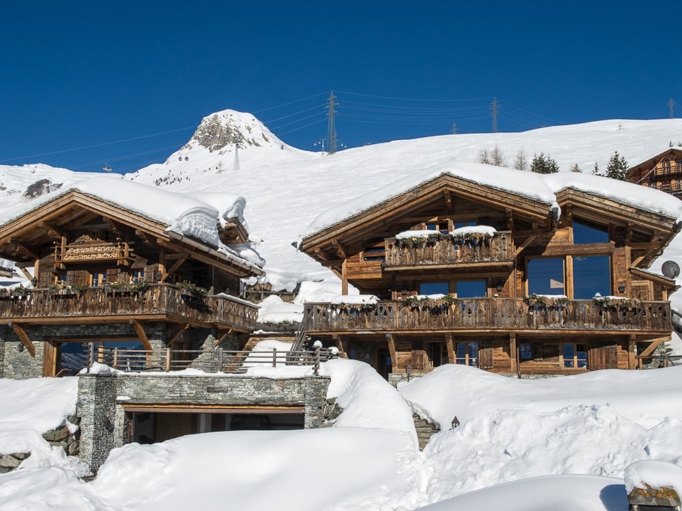 Luxurious wooden chalets are nestled in a snowy landscape with a clear blue sky overhead. These exquisite buildings feature large windows and balconies, perfect for luxury travel enthusiasts, and a snow-covered mountain peak rises majestically in the background. David Pearson Travel
