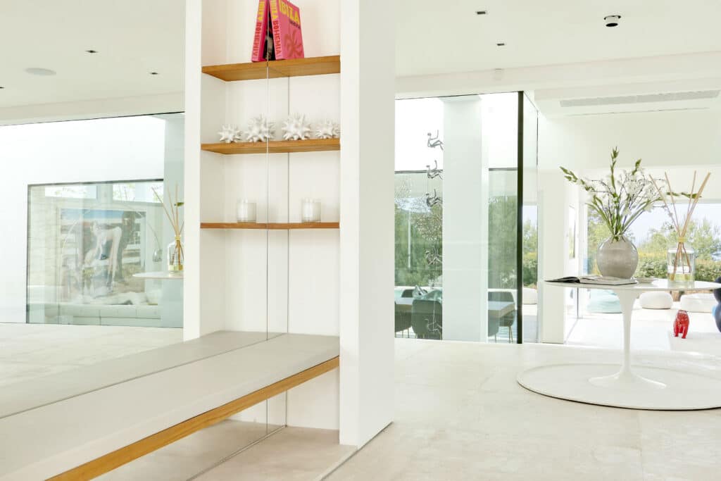 A modern, minimalist interior with white walls and large windows exudes an atmosphere akin to luxury travel. A narrow shelf holds books and decorative items, while a round table with a vase of flowers completes the look. Natural light enhances the airy ambiance. David Pearson Travel