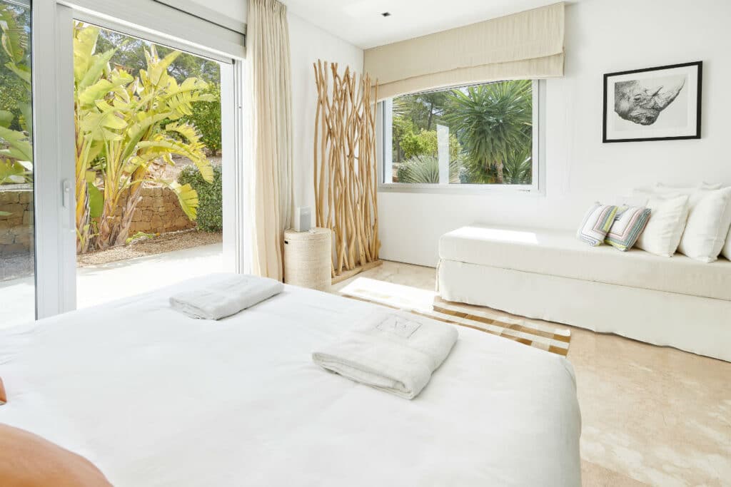 Experience a bright, luxurious bedroom featuring a large bed with crisp white linens. Enjoy the elegance of a white sofa adorned with decorative cushions by the window and tall plant décor. A glass door reveals lush garden greenery, complemented by tan marble flooring and framed art, perfect for luxury travel relaxation. David Pearson Travel