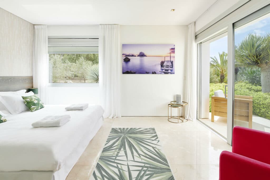 Bright, modern bedroom embodying luxury travel with a large bed and crisp white bedding. A tropical-themed rug graces the floor. The balcony offers a lush garden view, while a serene landscape photograph adorns the white wall. Sliding glass doors seamlessly lead to an outdoor escape. David Pearson Travel