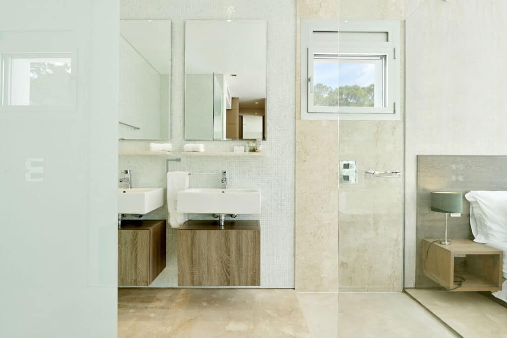 Modern bathroom with two white sinks on wooden vanities, large mirrors above each. A small window graces the beige-tiled wall. Glass shower enclosure and a glimpse of a bed in an adjoining room evoke luxury travel vibes. Neutral tones create a serene atmosphere. David Pearson Travel