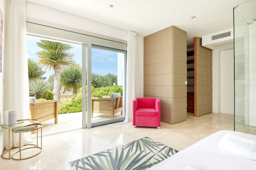 A bright, modern bedroom epitomizes luxury travel with a glass sliding door opening to a patio featuring two wooden chairs. Inside, a red armchair, small round table, and patterned rug complement the minimalist decor in neutral tones bathed in ample natural light. David Pearson Travel