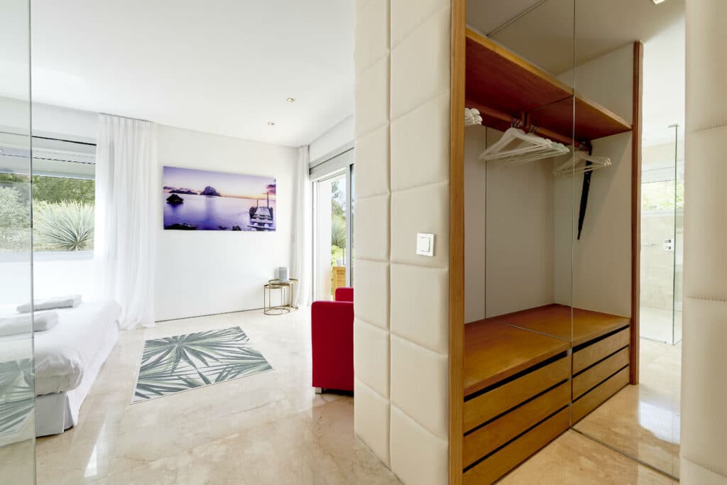 Experience luxury travel in our elegant hotel room, showcasing minimalist design. The space features a cozy bed, a large photo on the wall, and a glass wardrobe. Sunlight floods through expansive windows, illuminating the marble flooring while highlighting the vibrant red chair and leafy rug. David Pearson Travel