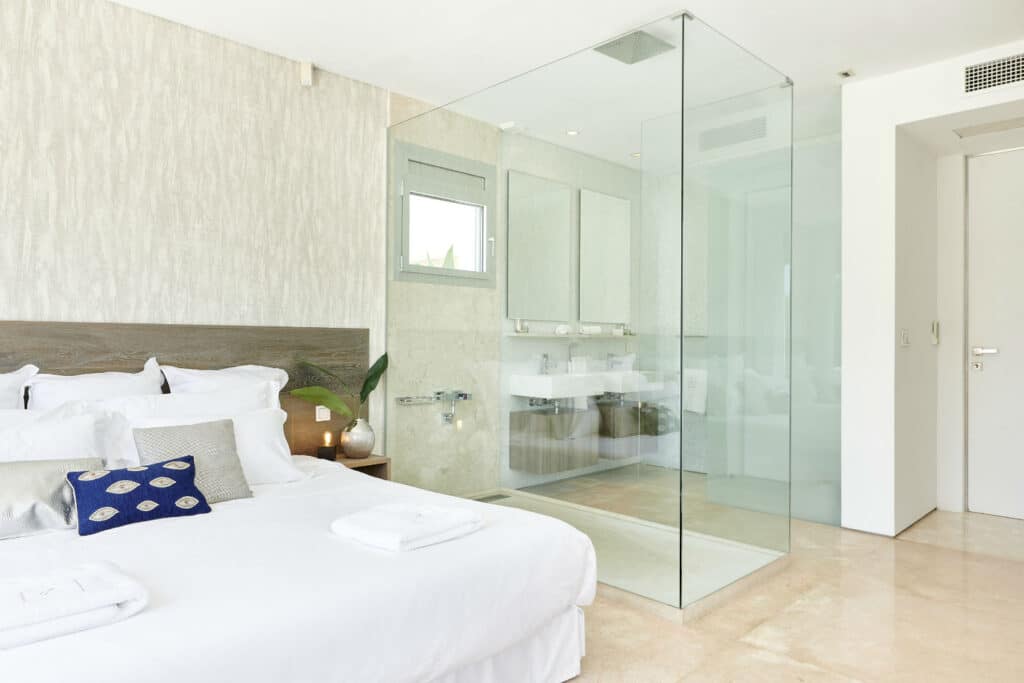 A modern bedroom designed for luxury travel enthusiasts, featuring a white bed with a wooden headboard and decorative pillows. A glass-enclosed shower in the corner complements the sink with a large mirror, set against beige flooring and light-colored walls. David Pearson Travel