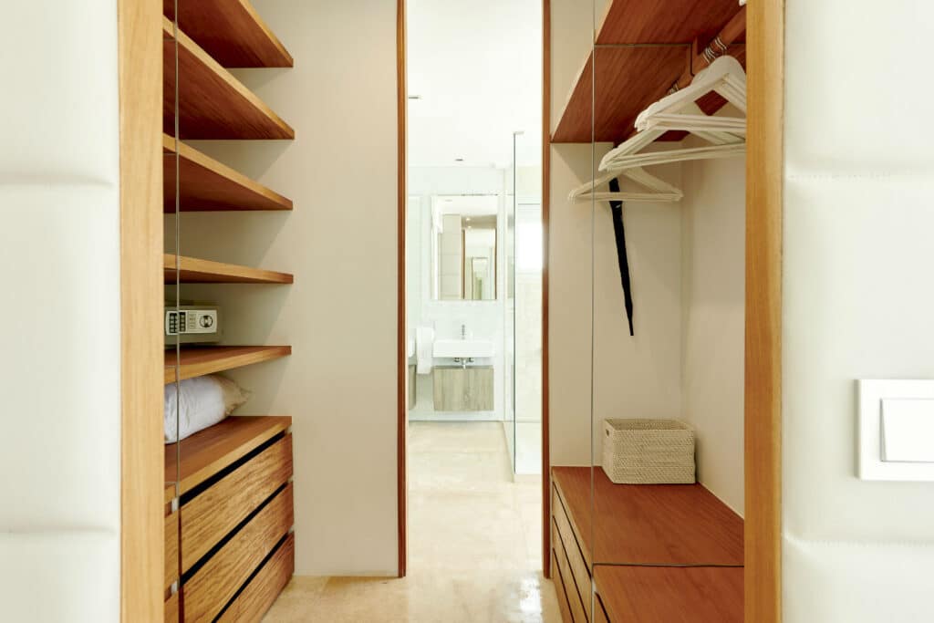 A walk-in closet with wooden shelves and drawers on either side showcases luxury travel essentials like hangers and a basket. A small safe and folded pillow are visible, while the closet leads to a well-lit bathroom with a mirror and sink in the background. David Pearson Travel