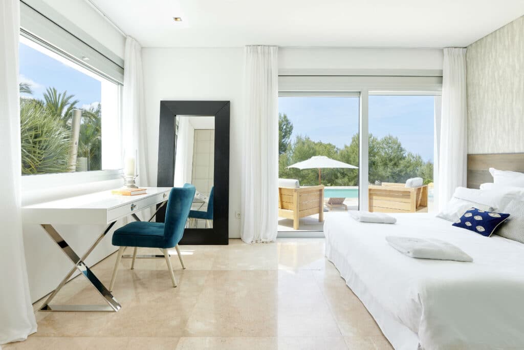 Bright, modern bedroom with a large bed and a hint of luxury travel flair. A blue chair by the white desk, full-length mirror, and floor-to-ceiling windows open to a patio. Outdoor seating and a pool shimmer invitingly amidst the trees under a clear sky. David Pearson Travel