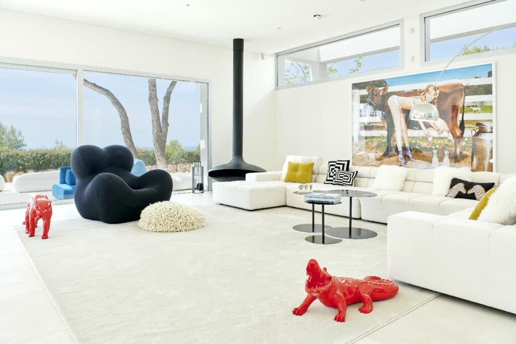 Modern living room with a white sectional sofa, black rounded chair, and red alligator sculptures evokes the essence of luxury travel. A wall-mounted fireplace and artwork depicting a pool scene add elegance, while large windows frame a view of trees. A beige rug adds warmth to the chic space. David Pearson Travel