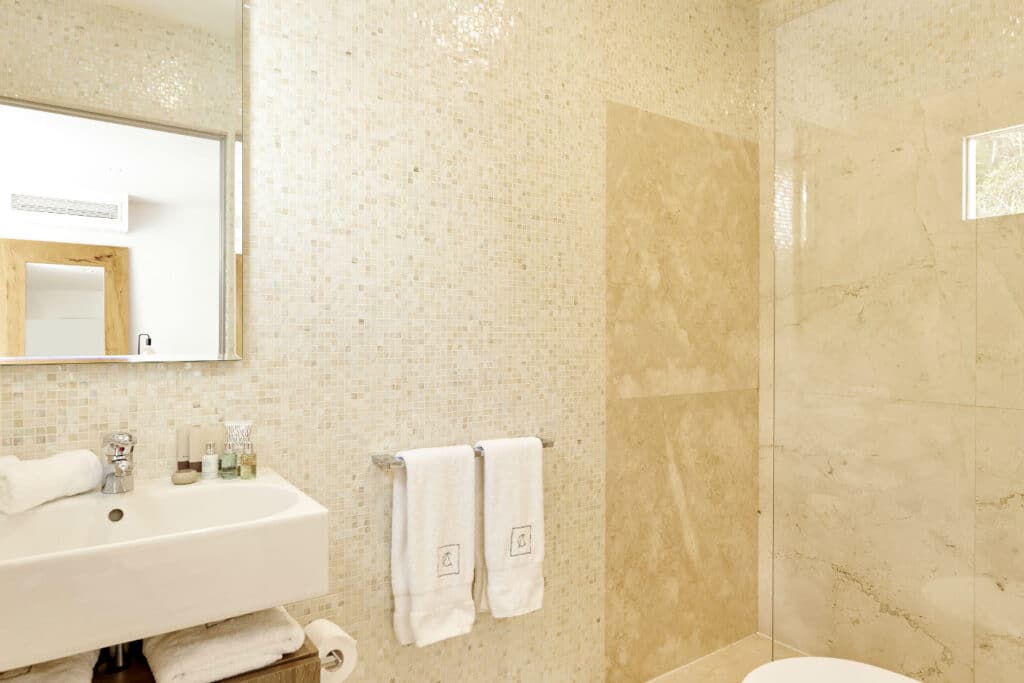 A modern bathroom with beige tiled walls and a glass-enclosed shower, perfect for those who appreciate the elegance of luxury travel. A white sink with a mirror above it sits on the left, while two plush white towels hang invitingly on a rack, complemented by neatly arranged toiletries. David Pearson Travel