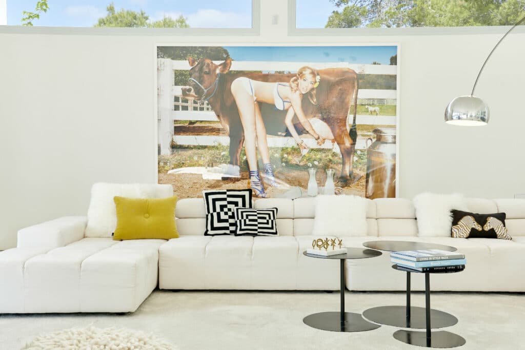 A modern living room exuding a luxury travel vibe features a large white sectional sofa adorned with patterned cushions. Above, captivating wall art depicts a person interacting with a cow. A sleek metallic floor lamp and round nesting tables with travel books complete the room. David Pearson Travel