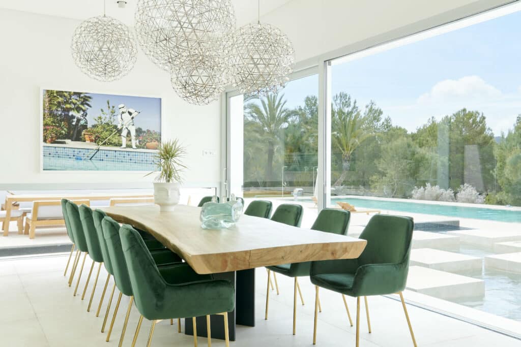 A modern dining room exudes luxury travel vibes with its long wooden table and plush green velvet chairs. Large spherical pendant lights hang overhead, while glass doors reveal a pool and lush greenery outside. A framed photo of an exotic poolside scene adds to the opulent atmosphere. David Pearson Travel