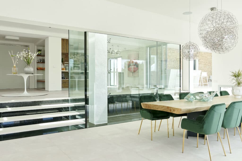 A modern dining area with a wooden table surrounded by green velvet chairs evokes the ambiance of luxury travel. Decorative wire pendant lights hang above, illuminating glass walls that divide the space, revealing another room with natural stone walls and large windows. David Pearson Travel