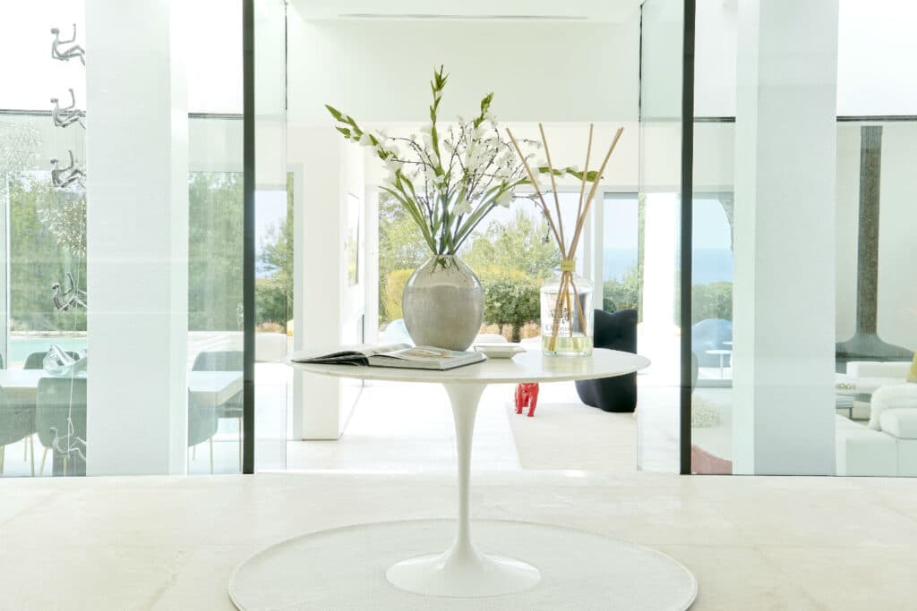 A minimalist, bright room evokes the essence of luxury travel, featuring a round white table with a gray vase containing white flowers and a small glass diffuser. Large glass doors open to an outdoor area with chic chairs and lush greenery, inviting you into a world of serene elegance. David Pearson Travel