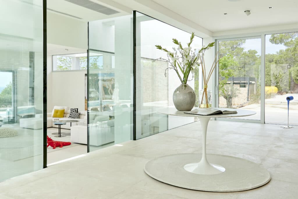 The modern interior of this luxurious home, akin to a luxury travel retreat, features expansive glass walls and doors. A white round table with a vase of white flowers and a book is centered in the room, while a stylish white sofa adorned with colorful cushions graces the background. David Pearson Travel