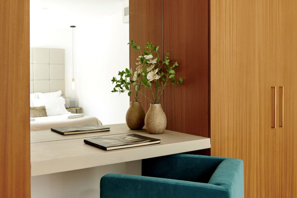 A modern bedroom exudes luxury travel vibes with a wooden vanity adorned by two vases holding greenery. Books rest on the countertop while a teal chair awaits underneath. The bed, draped in white linens and crowned by a padded headboard, hints at refined relaxation. David Pearson Travel