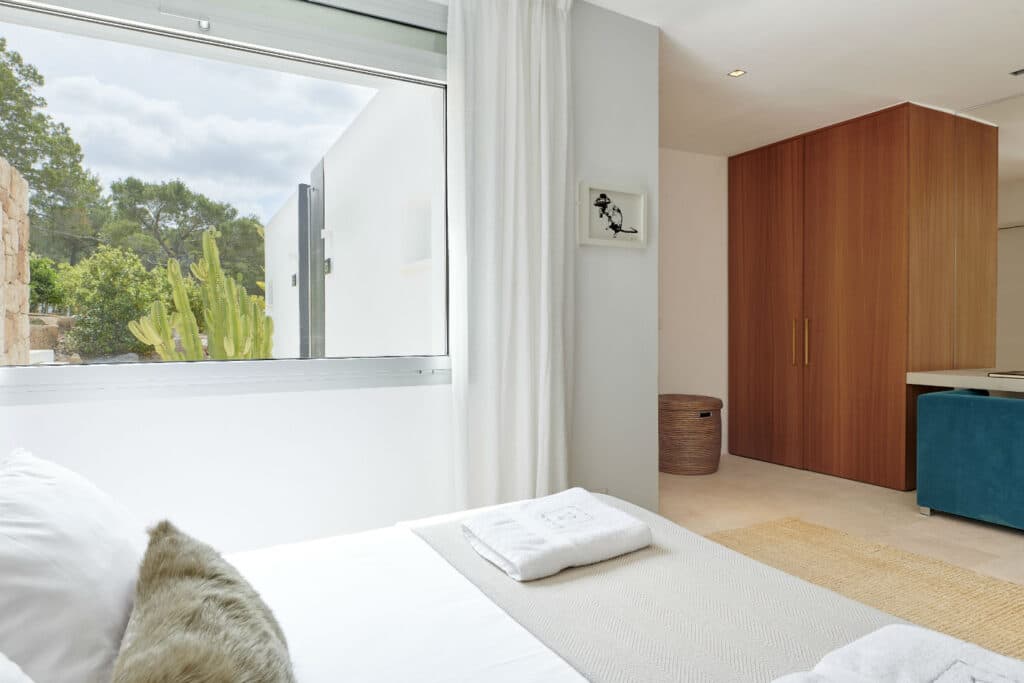 Modern bedroom with a large window, white walls, and light wooden furniture. A bed with white sheets and towels faces the window showing greenery outside—a touch of luxury travel found in the woven basket and minimalist art piece adorning the wall. David Pearson Travel