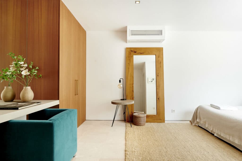 A minimalist bedroom evokes the serenity of luxury travel, featuring a large wooden-framed mirror, a wooden wardrobe, and a teal upholstered chair at a white desk. A round side table with a lamp complements the bed with white bedding on a woven rug, while a potted plant adds a touch of nature. David Pearson Travel