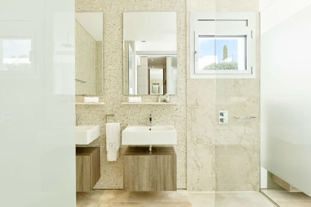 Experience a touch of luxury travel in this modern bathroom, adorned with beige mosaic tiles. It features dual sinks with elegant wooden cabinets, a large mirror, and a bright window. The glass shower enclosure and neutral-toned tiles enhance the rooms clean and sophisticated aesthetic. David Pearson Travel