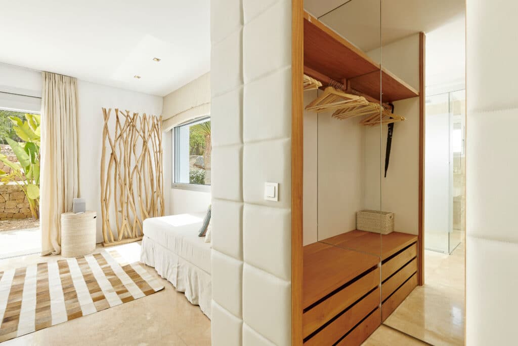 Bright, modern bedroom with natural light pouring in, perfect for luxury travel enthusiasts. Features a bed with a white cover, striped rug, wooden accent wall with branches, and an open closet with mirrors and hangers. A large window overlooks enchanting greenery. David Pearson Travel