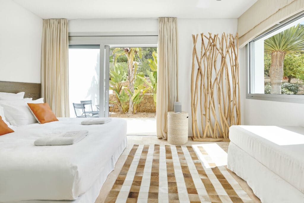 A bright, minimalist bedroom exudes luxury travel vibes with its large bed, crisp white linens, and vibrant orange pillows. A striped rug anchors the space as natural light pours in through glass doors, unveiling a lush garden. Tall wooden branches add an elegant touch in the corner. David Pearson Travel