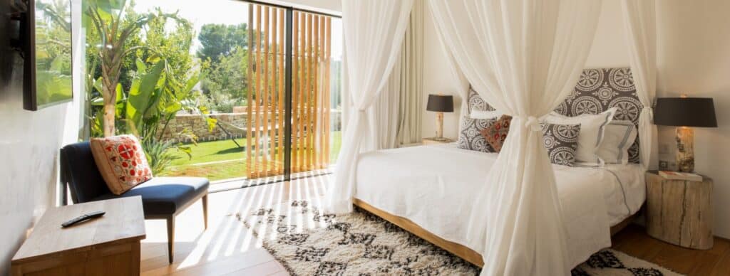 A bright, cozy bedroom with a canopy bed, decorative pillows, and stylish lamps offers a touch of luxury travel. Large glass doors with wooden details open to a lush garden. A chair with a cushion, a TV, and a patterned rug complement the serene atmosphere. David Pearson Travel