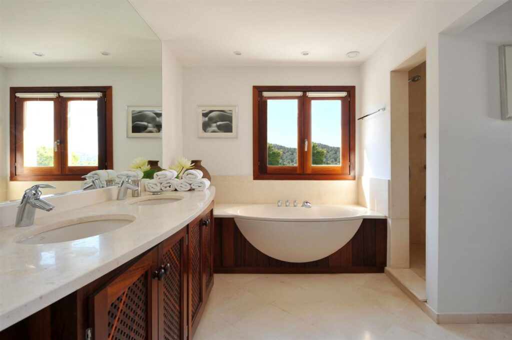 Modern bathroom with a large mirror reflecting double sink vanity, decorative plants, and neatly stacked towels. Reminiscent of luxury travel destinations, a bathtub sits beneath two large windows with scenic outdoor views. The room is adorned in neutral tones and wood accents. David Pearson Travel
