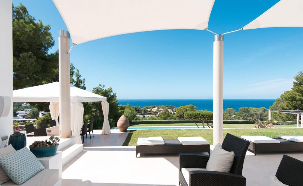 Experience luxury travel on a stunning outdoor patio featuring white shade sails and lounge chairs that overlook a lush lawn and the ocean. A large vase and trees frame the view, with a charming gazebo on the left under a clear blue sky. David Pearson Travel