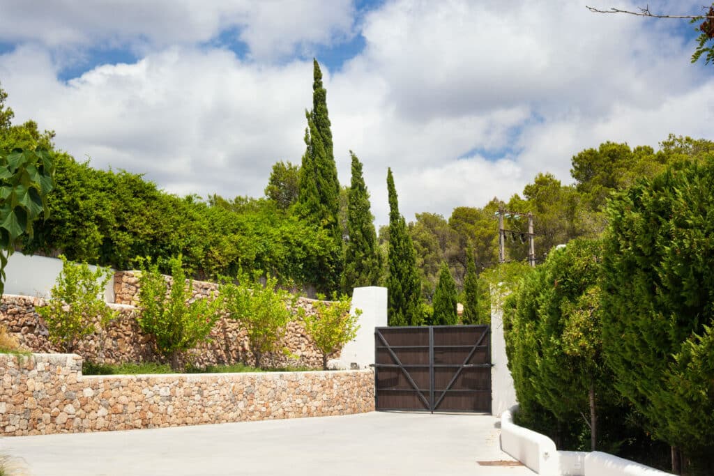 A tall wooden gate, evoking luxury travel, stands closed against a backdrop of lush greenery and stone walls. Tall cypress trees and a cloudy blue sky rise above the walls, adding to the opulent atmosphere. The smooth white driveway leading up to the gate enhances the scenes elegance. David Pearson Travel