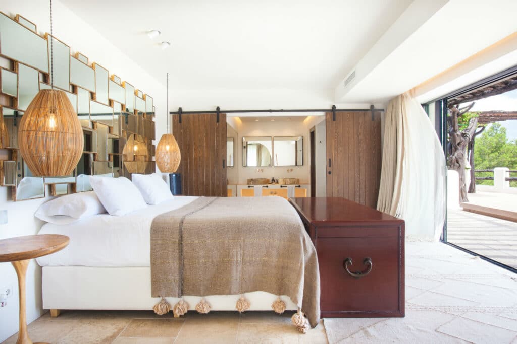 A spacious bedroom, perfect for luxury travel enthusiasts, features a large bed, wooden chest, and layered mirrors on the wall. Warm pendant lights hang above nightstands. An open sliding door reveals a balcony with lush greenery outside. The decor is modern and earthy. David Pearson Travel
